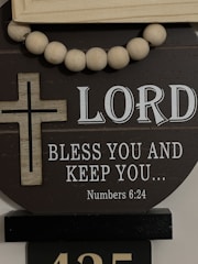 A decorative display featuring a wooden cross and a string of large wooden beads. The background has the text 'LORD BLESS YOU AND KEEP YOU... Numbers 6:24' in white, set against a dark circular board.
