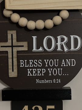 A decorative display featuring a wooden cross and a string of large wooden beads. The background has the text 'LORD BLESS YOU AND KEEP YOU... Numbers 6:24' in white, set against a dark circular board.