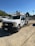 A white utility truck parked on a gravel surface with several other vehicles in the background. The truck has a logo on the side, a license plate, and a mounted rack on top. The setting appears to be an outdoor industrial area with a green hillside and trees in the background under a clear blue sky.