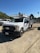 A white utility truck parked on a gravel surface with several other vehicles in the background. The truck has a logo on the side, a license plate, and a mounted rack on top. The setting appears to be an outdoor industrial area with a green hillside and trees in the background under a clear blue sky.