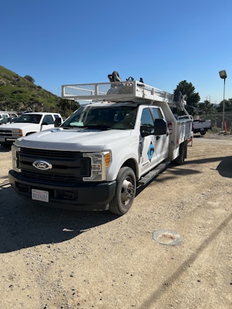A white utility truck parked on a gravel surface with several other vehicles in the background. The truck has a logo on the side, a license plate, and a mounted rack on top. The setting appears to be an outdoor industrial area with a green hillside and trees in the background under a clear blue sky.