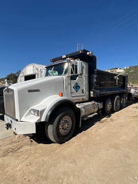 A large white and black dump truck is parked on a dirt road. The truck features a prominent cab with a glossy finish and several visible wheels. The backdrop includes a bright blue sky and some hills.
