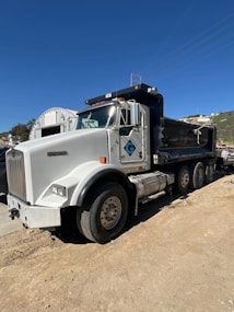 A large white and black dump truck is parked on a dirt road. The truck features a prominent cab with a glossy finish and several visible wheels. The backdrop includes a bright blue sky and some hills.