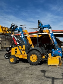 Heavy machinery including a yellow backhoe loader and a blue telehandler are parked on a concrete surface. The vehicles have distinct brand markings and are positioned under a clear sky with some power lines in the background.