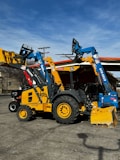 Heavy machinery including a yellow backhoe loader and a blue telehandler are parked on a concrete surface. The vehicles have distinct brand markings and are positioned under a clear sky with some power lines in the background.