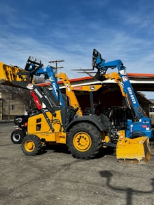 Heavy machinery including a yellow backhoe loader and a blue telehandler are parked on a concrete surface. The vehicles have distinct brand markings and are positioned under a clear sky with some power lines in the background.