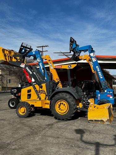 Heavy machinery including a yellow backhoe loader and a blue telehandler are parked on a concrete surface. The vehicles have distinct brand markings and are positioned under a clear sky with some power lines in the background.