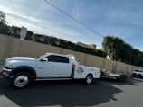 A large white pickup truck is parked on a street, attached to a flatbed trailer. The area is residential, with a high beige wall and green hedges along the sidewalk. Power lines run overhead, and there is a no parking sign nearby.