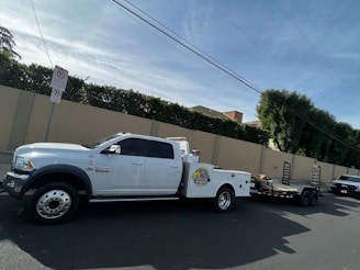 A large white pickup truck is parked on a street, attached to a flatbed trailer. The area is residential, with a high beige wall and green hedges along the sidewalk. Power lines run overhead, and there is a no parking sign nearby.