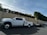 A large white pickup truck is parked on a street, attached to a flatbed trailer. The area is residential, with a high beige wall and green hedges along the sidewalk. Power lines run overhead, and there is a no parking sign nearby.
