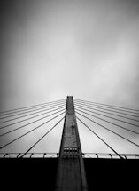 A black and white image featuring a bridge with a series of cables fanning out from a central pillar. The viewpoint is from below, creating a sense of upward perspective against a cloudy sky.