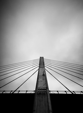 A black and white image featuring a bridge with a series of cables fanning out from a central pillar. The viewpoint is from below, creating a sense of upward perspective against a cloudy sky.