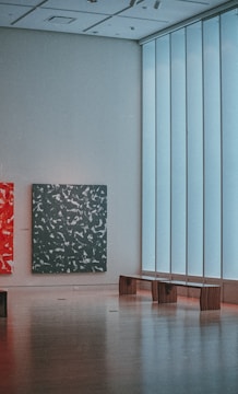 A modern art gallery interior with parquet flooring, featuring large, abstract paintings on the walls. One painting has a vibrant red background with white patterns, while another has a dark background with light patterns. The room has a minimalist design, with a large strip of diffused natural light coming through tall windows, and two wooden benches placed against the wall under the windows.