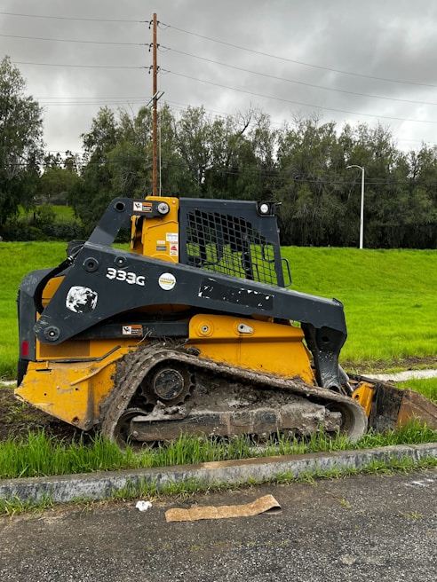A yellow and black skid-steer loader with muddy tracks is parked alongside a grassy area. Overhead, power lines run across the background against a cloudy, overcast sky. Behind the machinery, there are tall trees and utility poles.
