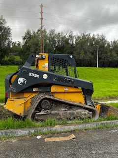 A yellow and black skid-steer loader with muddy tracks is parked alongside a grassy area. Overhead, power lines run across the background against a cloudy, overcast sky. Behind the machinery, there are tall trees and utility poles.