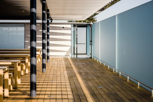 A modern outdoor space features elongated wooden decking and benches arranged in parallel under a slatted roof casting striped shadows. Black and white cylindrical poles support the structure, with blue-tinted glass walls creating a private, serene atmosphere. A door enclosed by glass panels is at one end of the walkway.
