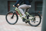 A person is riding a light green electric bicycle with the brand name 'Levit' visible on the frame. The cyclist is dressed casually in a dark top and light gray pants, moving swiftly on a paved surface. The background is slightly blurred, suggesting motion.