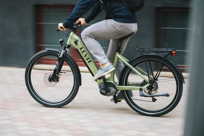 A person is riding a light green electric bicycle with the brand name 'Levit' visible on the frame. The cyclist is dressed casually in a dark top and light gray pants, moving swiftly on a paved surface. The background is slightly blurred, suggesting motion.