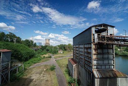 An industrial scene with large metal structures and a pathway leading towards a cooling tower in the background. The landscape is interspersed with greenery, trees, and a river or water body running beside the metal buildings. The sky is bright and partly cloudy, adding to the contrast between the industrial architecture and natural elements.