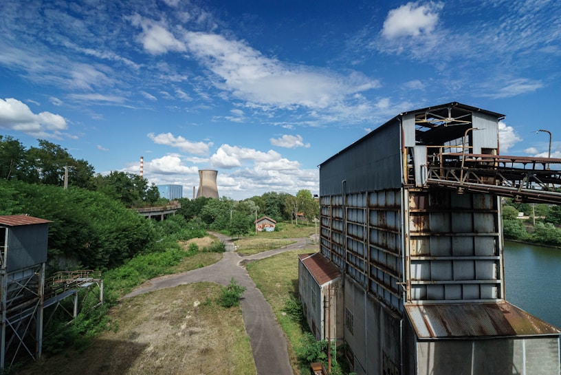 An industrial scene with large metal structures and a pathway leading towards a cooling tower in the background. The landscape is interspersed with greenery, trees, and a river or water body running beside the metal buildings. The sky is bright and partly cloudy, adding to the contrast between the industrial architecture and natural elements.