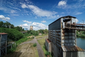 An industrial scene with large metal structures and a pathway leading towards a cooling tower in the background. The landscape is interspersed with greenery, trees, and a river or water body running beside the metal buildings. The sky is bright and partly cloudy, adding to the contrast between the industrial architecture and natural elements.