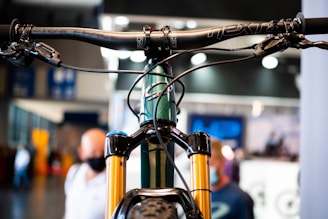 Close-up view of a bicycle handlebar and front suspension fork. The focus is on the intricate details of the bike components, showing the handlebars with brake levers and cables. The background is slightly blurred with people in an indoor setting, perhaps at an exhibition or store.