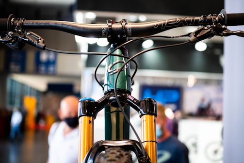 Close-up view of a bicycle handlebar and front suspension fork. The focus is on the intricate details of the bike components, showing the handlebars with brake levers and cables. The background is slightly blurred with people in an indoor setting, perhaps at an exhibition or store.