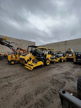 A collection of large construction vehicles and equipment parked in a yard against a backdrop of a cloudy sky and concrete walls. The machinery includes several yellow rollers, a backhoe, and other types of construction equipment.