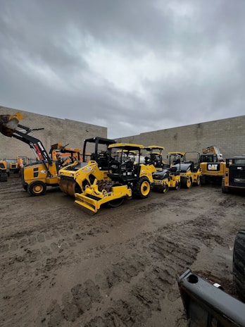 A collection of large construction vehicles and equipment parked in a yard against a backdrop of a cloudy sky and concrete walls. The machinery includes several yellow rollers, a backhoe, and other types of construction equipment.