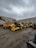 A collection of large construction vehicles and equipment parked in a yard against a backdrop of a cloudy sky and concrete walls. The machinery includes several yellow rollers, a backhoe, and other types of construction equipment.