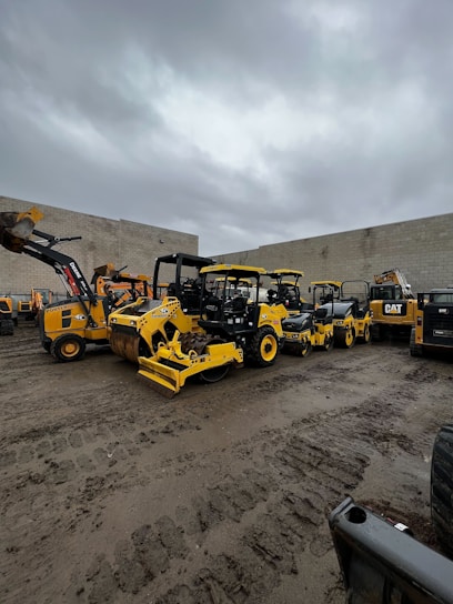 Construction equipment and machinery lined up at a rental yard on a sunny day.