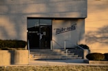 The image displays the entrance of a brick building featuring a black glass door with the address '333 LEO ST.' The building has a cream-colored brick exterior with 'Mike-sell's' logo in blue. In front of the entrance, there are concrete steps with metal railings, and landscaped bushes and grass.