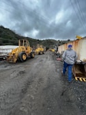 Two yellow construction vehicles are parked on a muddy road, surrounded by hilly terrain and cloudy skies. A person in a gray jacket and blue pants walks away from the camera towards the machinery. On the right, there are shipping containers and construction materials, while overhead power lines stretch across the sky.