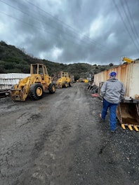 Two yellow construction vehicles are parked on a muddy road, surrounded by hilly terrain and cloudy skies. A person in a gray jacket and blue pants walks away from the camera towards the machinery. On the right, there are shipping containers and construction materials, while overhead power lines stretch across the sky.