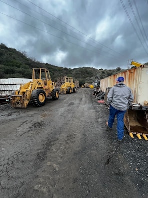 Two yellow construction vehicles are parked on a muddy road, surrounded by hilly terrain and cloudy skies. A person in a gray jacket and blue pants walks away from the camera towards the machinery. On the right, there are shipping containers and construction materials, while overhead power lines stretch across the sky.
