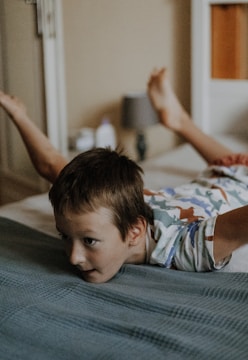 A young child is lying face down on a bed with their arms and legs extended outward. The child is wearing a colorful shirt with a pattern and appears to be in a playful pose, possibly pretending to fly like an airplane. The room is softly lit, and there are various home items in the background, including a lamp and a bottle.