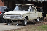A vintage car is elevated on jacks in front of a garage, showing signs of rust and age. The vehicle lacks tires and has parts stored on its roof. It is placed beside a modern black car, with various tools and items visible in the surrounding garage area.