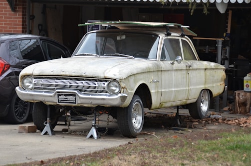 A vintage car is elevated on jacks in front of a garage, showing signs of rust and age. The vehicle lacks tires and has parts stored on its roof. It is placed beside a modern black car, with various tools and items visible in the surrounding garage area.