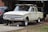 A vintage car is elevated on jacks in front of a garage, showing signs of rust and age. The vehicle lacks tires and has parts stored on its roof. It is placed beside a modern black car, with various tools and items visible in the surrounding garage area.