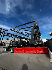 Several large construction lifts are parked in an outdoor lot, each with long extendable arms. Bright red barricades are placed in front of the machinery. The sky above is a clear, vivid blue with some wispy clouds.