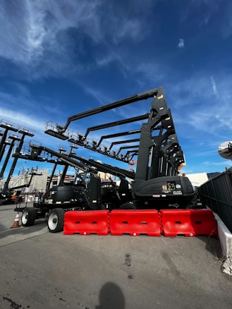 Several large construction lifts are parked in an outdoor lot, each with long extendable arms. Bright red barricades are placed in front of the machinery. The sky above is a clear, vivid blue with some wispy clouds.