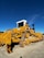 A large yellow bulldozer with caterpillar tracks sits on a dirt surface under a clear blue sky. The machinery is robust, with a white cab on top and heavy-duty metal components visible on the sides and front.