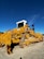 A large yellow bulldozer with caterpillar tracks sits on a dirt surface under a clear blue sky. The machinery is robust, with a white cab on top and heavy-duty metal components visible on the sides and front.