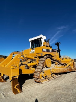 A large yellow bulldozer with caterpillar tracks sits on a dirt surface under a clear blue sky. The machinery is robust, with a white cab on top and heavy-duty metal components visible on the sides and front.