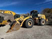 A large yellow construction vehicle, specifically a wheel loader, is parked outdoors on a gravel surface. Accompanying the loader is an excavator with a large bucket attachment. The background consists of dry, scrubby hills under a clear blue sky.