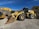 A large yellow construction vehicle, specifically a wheel loader, is parked outdoors on a gravel surface. Accompanying the loader is an excavator with a large bucket attachment. The background consists of dry, scrubby hills under a clear blue sky.