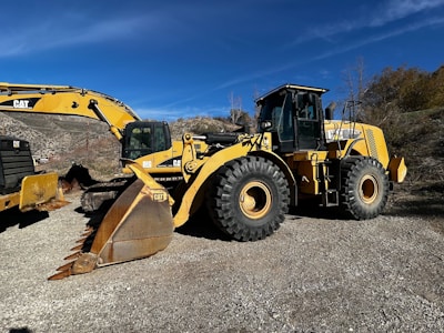 A large yellow construction vehicle, specifically a wheel loader, is parked outdoors on a gravel surface. Accompanying the loader is an excavator with a large bucket attachment. The background consists of dry, scrubby hills under a clear blue sky.