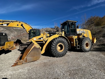 A large yellow construction vehicle, specifically a wheel loader, is parked outdoors on a gravel surface. Accompanying the loader is an excavator with a large bucket attachment. The background consists of dry, scrubby hills under a clear blue sky.
