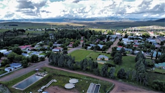 Aerial view of a small town with a combination of residential buildings, trees, roads, and some open green spaces. In the foreground, there are some industrial structures perhaps related to water treatment, alongside ponds or reservoirs. The town is surrounded by lush green fields and a range of hills or mountains can be seen in the background under a partly cloudy sky.