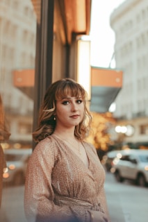A woman with wavy, shoulder-length hair stands confidently on a city street. She wears a patterned dress and subtle makeup. The street is bustling with cars and illuminated by warm, soft lights reflecting off tall buildings.